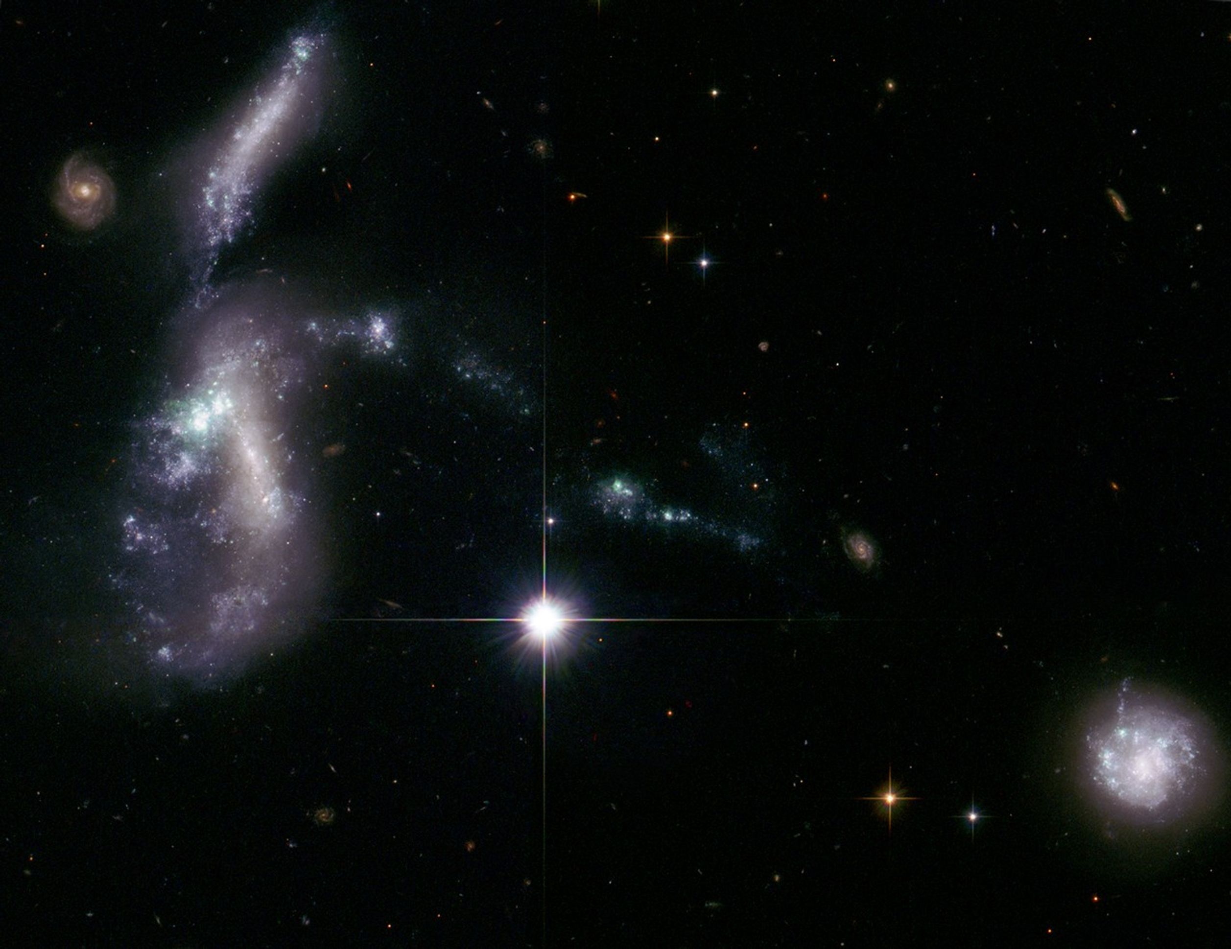The stars, gas, and dust seem to form a blue, white, and purple knot-shaped bow on the left, with a small red-orange spiral in the background in the upper left. A bright white foreground star just lower-left of center. Another smaller, purple-white spiral in the bottom-right.